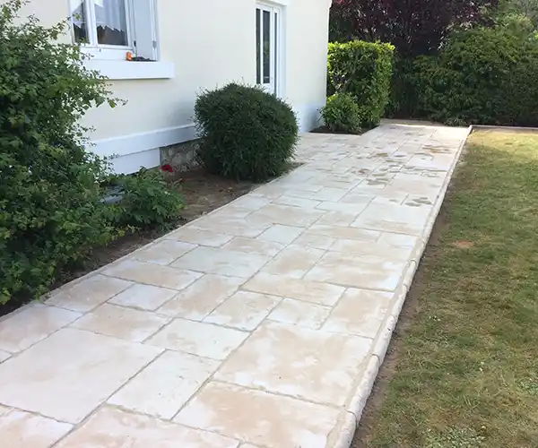 Une allée pavée longe une maison blanche, bordée de buissons verts et d&#039;une pelouse. Les pavés clairs, disposés avec soin, agrémentent le jardin, idéal pour la création d&#039;une terrasse élégante à l&#039;arrière.