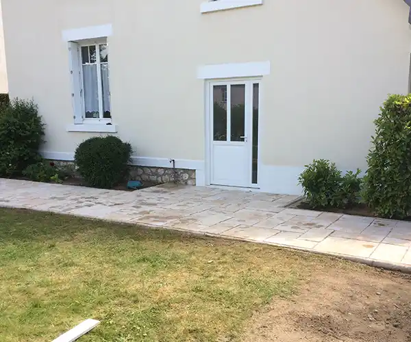 Une maison beige avec une fenêtre à cadre blanc et une porte vitrée s&#039;ouvre sur un patio en pierre claire, featuring la création d&#039;une terrasse en dallage pierre reconstituée. Des buissons verts bordent le mur, tandis que la pelouse et le sol nu se trouvent au premier plan.