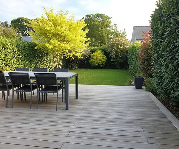 Une terrasse moderne en bois, un exemple étonnant de création terrasse en bois à Blois, comprend une table et quatre chaises donnant sur une pelouse verte bordée de haies, d&#039;arbres et d&#039;arbustes luxuriants dans le jardin de l&#039;arrière-cour.