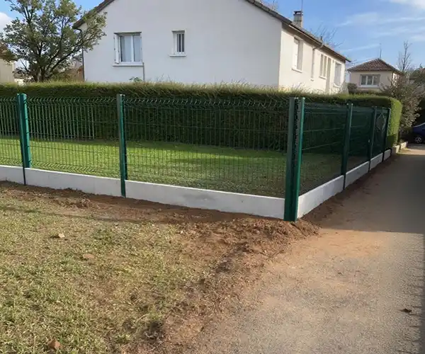 Une clôture métallique verte avec des panneaux en maille offre une pose de clôture rigide sur muret béton, entourant une haie bien taillée et une cour gazonnée devant une maison blanche, à côté d&#039;un sentier pavé et d&#039;une autre maison à l&#039;arrière-plan.