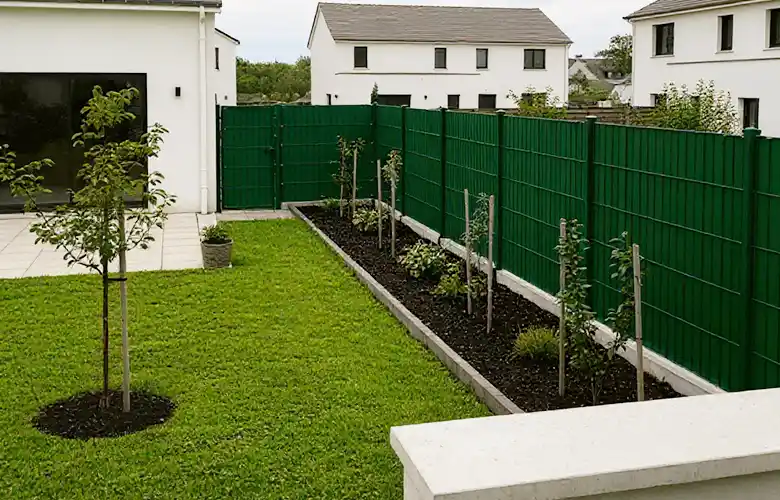 Une arrière-cour moderne avec une pelouse verte, de jeunes arbres, une plate-bande rectangulaire avec de petites plantes et une clôture métallique verte - un type de clôture élégant - encadrant des maisons blanches à l&#039;arrière-plan.