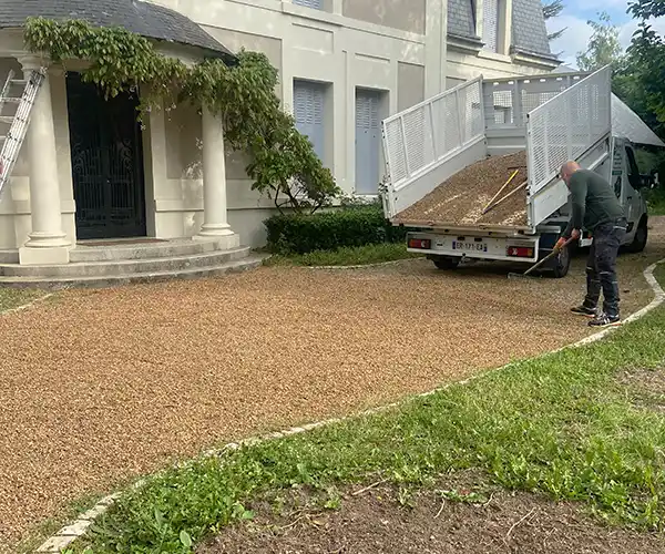 Un ouvrier répand du gravier sur une allée devant une maison, à l&#039;aide d&#039;un râteau, tandis qu&#039;un camion décharge du gravier à proximité. Cette réalisation d&#039;une allée en gravier stabilisé est encadrée par des marches menant à l&#039;entrée et par de la verdure bordant l&#039;allée.