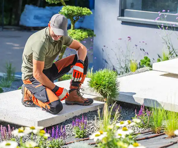 Un paysagiste est agenouillé sur un chemin de pierre, portant des gants de travail, une casquette et un pantalon de protection, tout en effectuant des travaux paysagers et en s'occupant de plantes et de fleurs à côté d'un bâtiment moderne.