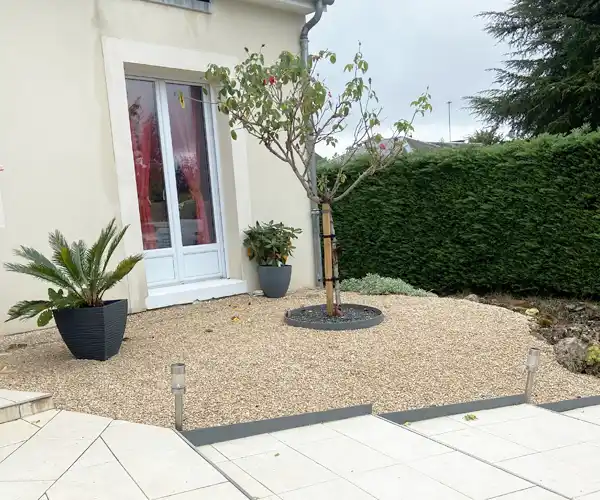 Un petit jardin avec un sol en gravier présente la création d'un massif autour d'un arbre central, des plantes en pot de chaque côté près d'une porte blanche, et une haie à l'arrière-plan. Le patio à l'avant est pavé de carreaux de couleur claire.