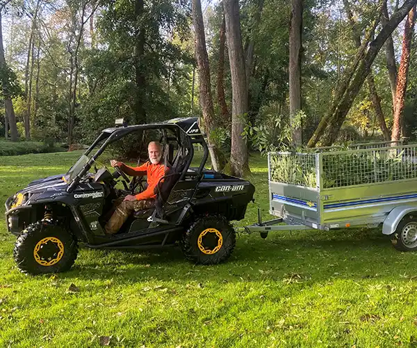 Michel GARAND conduit un buggy noir, tirant une petite remorque remplie de branches d'arbres taillées - preuve de récents travaux d'élagage - à travers une zone herbeuse entourée d'arbres.